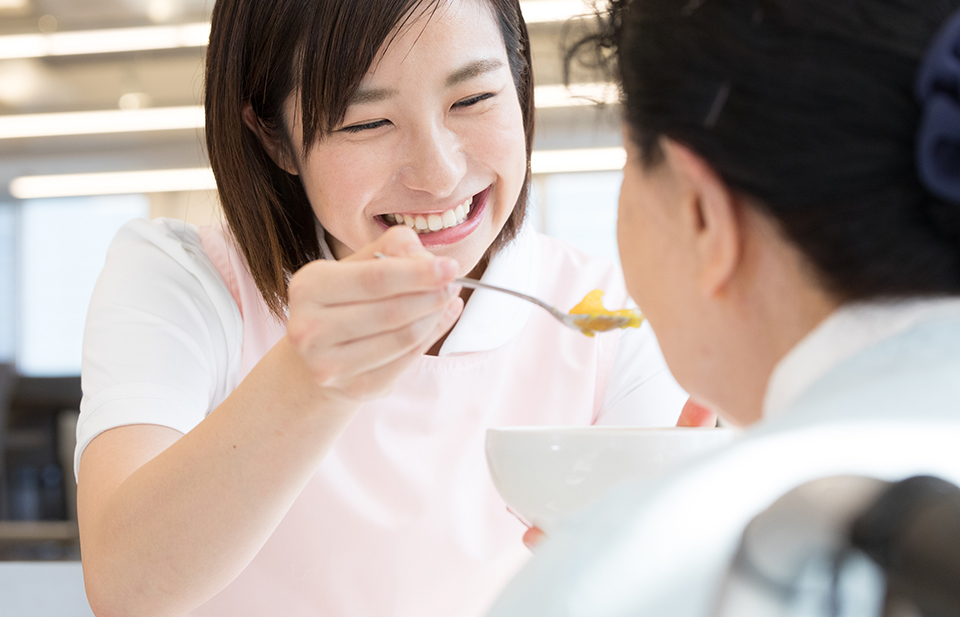 介護職員が入居者に食事の援助をしている風景。笑顔で対応している様子が見られる。